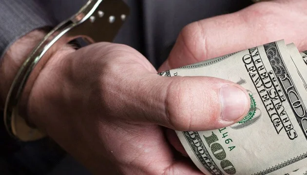 Рука в наручниках тримає стодоларові купюри, символізуючи хабар у $100 тис. посадовцю Укрзалізниці